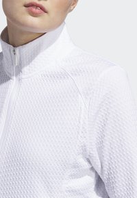 Casaco branco com fecho de correr, apresentando um tecido texturizado com um padrão de diamante, gola alta e detalhe de fecho suave.
