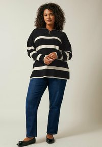 Schwarz-creme gestreifter Strickpullover mit einem Reißverschlusskragen, kombiniert mit dunkelblauen geraden Jeans und schwarzen Ballerinas.