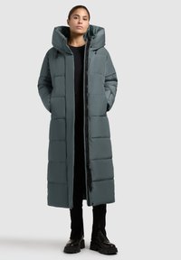 Long cappotto puffo in verde scuro con cappuccio, dotato di un design trapuntato, chiusura frontale con zip e tasche laterali. Tessuto liscio e isolante.