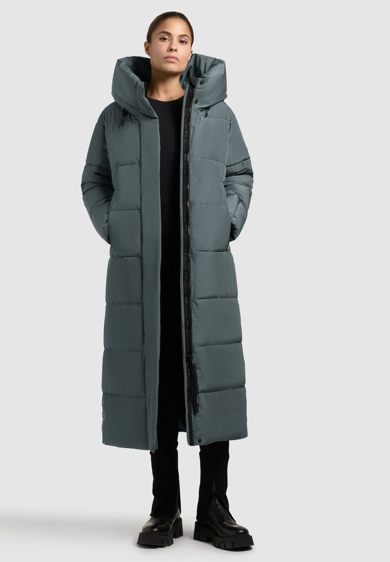 Long cappotto puffo in verde scuro con cappuccio, dotato di un design trapuntato, chiusura frontale con zip e tasche laterali. Tessuto liscio e isolante.