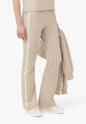 Onderlichaam van een persoon die beige patroon wijde broek met witte zijstrepen en bijpassende top draagt, houdend een beige jas en witte sneakers.