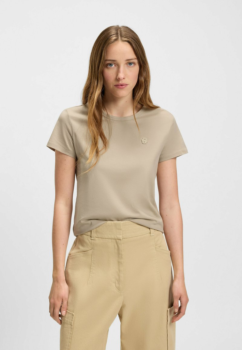 Beżowy t-shirt z krótkim rękawem z miękkiego materiału, z okrągłym dekoltem, z małym wytłoczonym logo na klatce piersiowej.