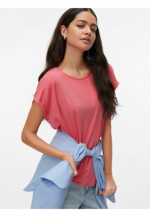 Woman with long dark hair wearing coral short-sleeve top and light blue jacket tied around waist, standing against white background.