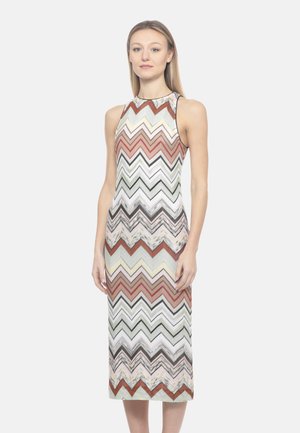 Donna che indossa un vestito senza maniche lungo fino alla caviglia con motivo a zigzag multicolore in tonalità di bianco, marrone, rosso e grigio, in piedi davanti a uno sfondo semplice.