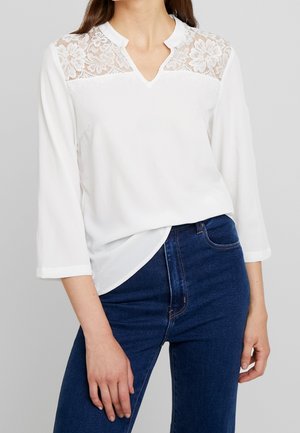 Kobieta w białej bluzce z długim rękawem z koronkowym kwiatowym zdobieniem na ramionach i ciemnoniebieskich dżinsach z wysokim stanem.