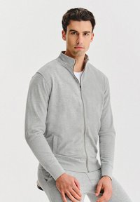 Jasnoszara bluza rozpinana na zamek z wysokim kołnierzem, długimi rękawami i przednimi kieszeniami. Wykonana z miękkiego materiału o gładkiej fakturze.