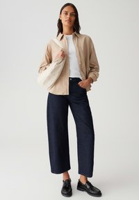 Vrouw in een beige overhemd over een witte top, met donkere wijde jeans, zwarte schoenen en een witte schoudertas, staande tegen een effen achtergrond.