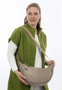 Borsa a tracolla beige con una forma curva, texture liscia e un accento decorativo a catena. Indossata sopra un capo verde a texture.