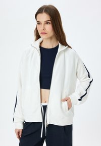 Haut de sweat à capuche blanc zippé avec des rayures latérales bleu marine, en tissu doux et avec des poches latérales ; présente un col haut et une coupe décontractée, porté sur un haut bleu marine.