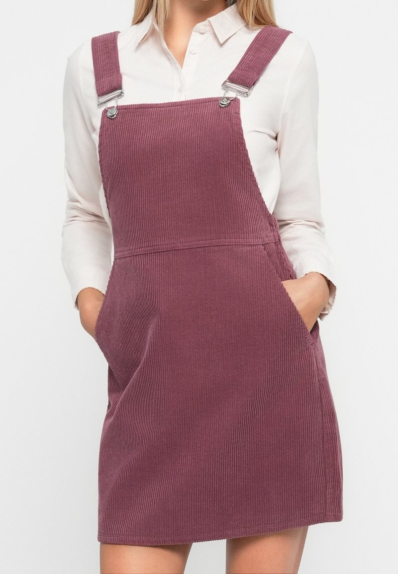 Femme portant une robe salopette en velours côtelé mauve avec des bretelles ajustables, sur une chemise blanche boutonnée, les mains dans les poches de la robe.