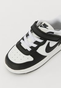 Baskets en cuir noir et blanc avec une boîte à orteils perforée, lacets blancs, sangle sur la languette et un logo Swoosh noir sur le côté.