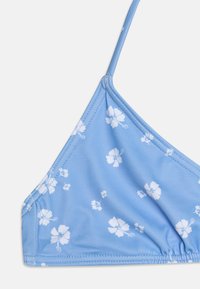 Světle modrý bikiny top s bílými květinovými vzory, trojúhelníkový tvar, tenké nastavitelné ramínka a nařasená detail na spodní části.