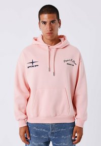 Jonge man in een lichtroze hoodie met een voorzak en logo's, gecombineerd met blauwe jeans met een herhalend patroon van witte tekst.