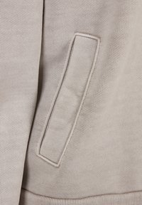 Tessuto di felpa beige con una texture morbida, dotato di una tasca laterale visibile con bordo cucito. Inquadratura ravvicinata che evidenzia i dettagli.