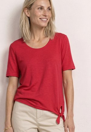 Femme souriante, portant un t-shirt rouge à manches courtes avec un nœud sur le côté, associé à un pantalon beige, debout contre un mur de couleur claire.