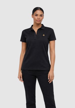 Donna in piedi che indossa una polo nera a maniche corte con un piccolo emblema sul petto e pantaloni neri su uno sfondo grigio uniforme.