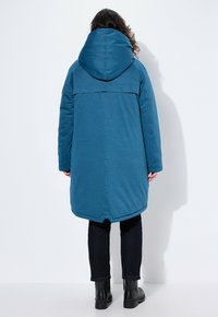 Turkusowa kurtka puchowa z dużym kapturem, posiada bezszwowy design, do kolan, noszona z czarnymi spodniami i butami.