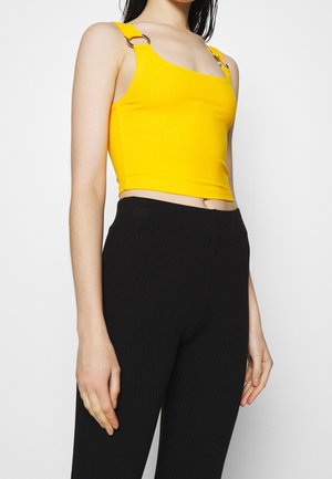 Gelbes geripptes Crop-Top mit diagonellem Ausschnitt und Metallringakzenten an den Trägern, kombiniert mit hoch geschnittenen schwarzen gerippten Hosen.