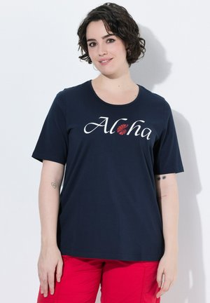 Kobieta o krótkich ciemnych kręconych włosach, ubrana w granatowy T-shirt z napisem "Aloha" i czerwone spodnie, stojąca na białym tle.