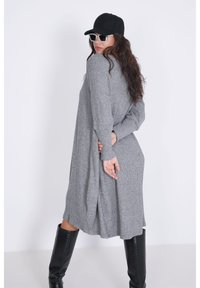 Lange grijze gebreide jurk met een losse pasvorm, zijsplitten, lange mouwen en ribbelstructuur. Gecombineerd met zwarte kniehoge laarzen en accessoires.