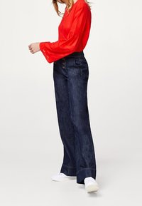 Mørkeblå denimjeans med bred benpasform, høj talje og synlige stikninger. Sammen med en lys rød langærmet top og hvide sneakers.