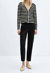 Donna vestita con un cardigan a righe bianche e nere con bottoni, pantaloni neri su misura e décolleté beige a punta bassa, in piedi su un pavimento bianco.