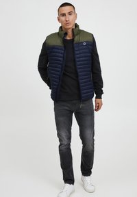 Tamsiai mėlyna ir alyvuogių žalia siūta liemenė su aukštu apykakle, užtrauktuku ir dviem šoninėmis kišenėmis, dėvima ant juodos palaidinės ir pilkų džinsų. Balti sportbačiai.