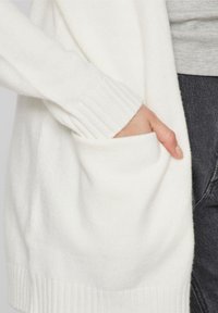 Cárdigan de punto blanco con puños acanalados, que presenta un bolsillo frontal. El material parece suave y texturizado, enfatizando un diseño acogedor.