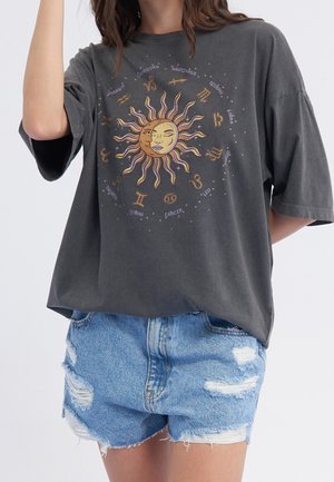 Femme portant un t-shirt gris oversize avec un motif soleil et signes du zodiaque, associé à un short en jean bleu déchiré.