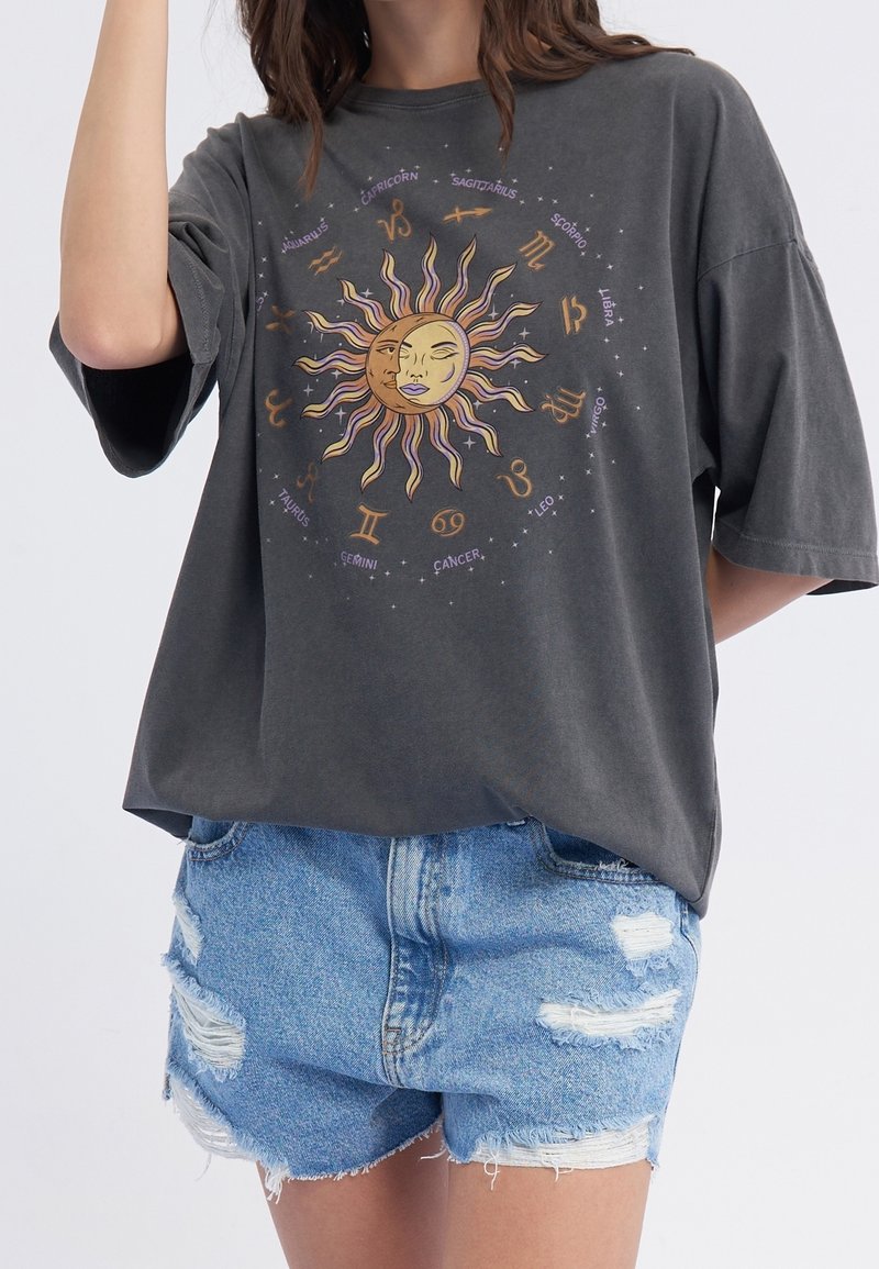 Femme portant un t-shirt gris oversize avec un motif soleil et signes du zodiaque, associé à un short en jean bleu déchiré.