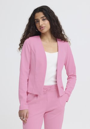 Femme aux longs cheveux bouclés portant un blazer et un pantalon roses avec un haut blanc, une main dans la poche, posant devant un fond uni.