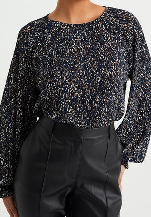 Persoon draagt een blauw, wit en bruin gespikkelde blouse met lange mouwen, ingestopt in een hoge zwarte leren broek met zijzakken.