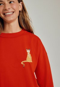 Hanorac roșu cu un imprimeu de leopard purtând ochelari de soare și o coroană florală pe pieptul sting. Material moale, design cu gât rotund.