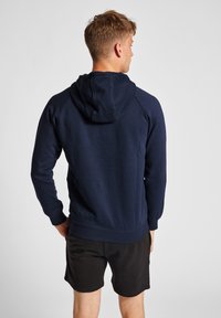 Marineblaue Kapuzenjacke mit Kordelzug, gerippten Bündchen und Saum. Aus weichem Stoff mit glatter Textur, getragen mit schwarzen Shorts.