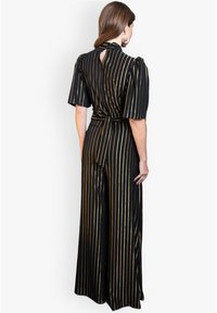 Gestreifter Jumpsuit in Schwarz und Gold mit kurzen Puffärmeln, Rückenknoten-Detail und offener Rückseite. Weite Hosenbeine, die bis zum Boden reichen.