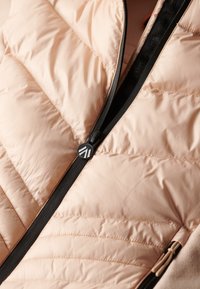 Lyserød polstret jakke med vandret quiltning, sort lynlåsdetalje og en rund sort logo-knap ved halsen. Blødt, letvægtsstof.