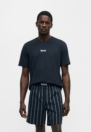 Mężczyzna ubrany w granatowy t-shirt BOSS i czarno-białe pionowo paskowane spodenki, stojący na tle jednolitego jasnego tła.