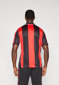 Αθλητική φανέλα με μαύρες και κόκκινες ρίγες, μικρό γιακά, κοντά μανίκια και το κείμενο "ISSA NISSA" στην πλάτη. Λείο, ελαφρύ ύφασμα.