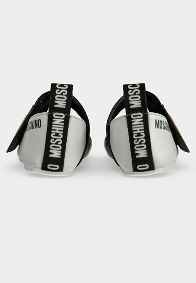 Двойка бели обувки без връзки с черни дръжки с текст "MOSCHINO", показани отзад на бял фон.