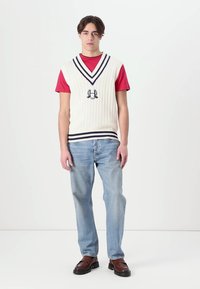 Strikket kremfarget vest med marineblå striper over en rød t-skjorte; lyseblå jeans og brune sko. Har et logo på brystet og ribbestruktur.