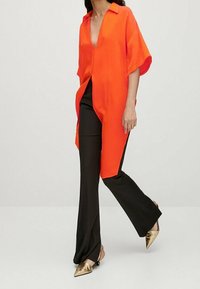 Tunique orange légère à manches courtes avec un profond décolleté, associée à un pantalon flare noir et des escarpins dorés à bout pointu.