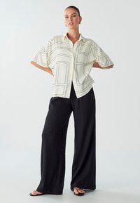 Mujer de pie con las manos en las caderas, vistiendo una camisa de botones de patrón crema, pantalones de pierna ancha negros, aretes de aro dorados y sandalias negras.