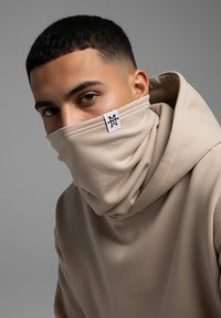 Beige hoodie with a high collar, featuring a logo tag. Fabric appears soft and smooth, draping closely around the face.