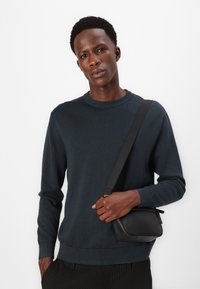 Dunkelblauer Strickpullover mit runden Ausschnitt und gerippten Bündchen, getragen mit einer kleinen schwarzen Umhängetasche. Glatte Textur, taillierte Passform.