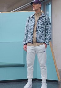 Jachetă cu vată pentru bărbați, cu un model floral în nuanțe de albastru și mov, cămașă bej, pantaloni sport albi și adidași albi. Șapcă purtată pe cap.