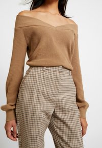 Sweter dziergany z dekoltem odkrywającym ramiona w kolorze wielbłądzim, zestawiony z brązowo-beżowymi, wysokimi, kraciastymi spodniami o dopasowanym kroju i subtelnej fakturze.