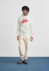 Ανοιχτό κρεμ φούτερ με κόκκινο logo "Just Do It", σετ με τζόγκερ στο ίδιο χρώμα και μαύρα αθλητικά παπούτσια. Λείο ύφασμα, άνετη εφαρμογή.