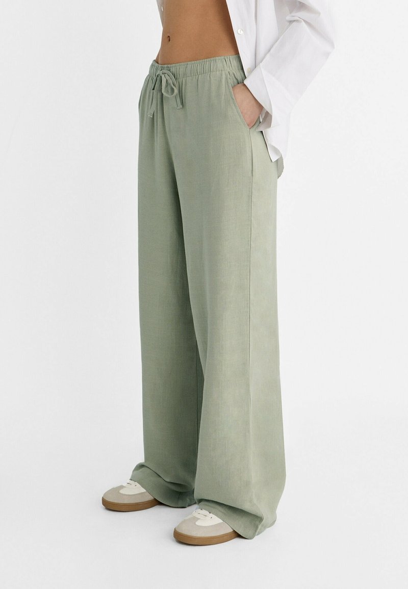 Personne portant un pantalon ample vert clair à cordon avec poches, associé à des baskets beige et blanches et une chemise blanche partiellement ouverte.