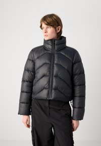 Casaco puffer preto com colarinho alto, apresentando um padrão em ziguezague, fecho de zip e punhos elásticos. Usado sobre calças cargo escuras.
