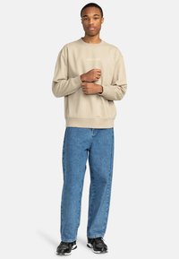 Beige bomullströja med rundad hals och en liten logotyp. Kombinerad med lösa blå denimjeans och svarta sneakers. Klassisk och avslappnad stil.